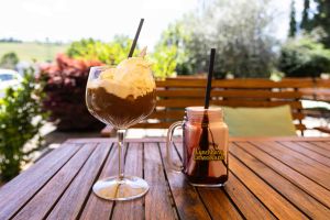 Ein Glas Affogato mit Sahne und Waffel steht neben einem Henkelglas mit rosa Milchshake auf einem hölzernen Terrassentisch.