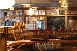 Blick über eine metallene Zapfanlage auf eine schwarze Wandtafel mit der Kreideaufschrift BILLARD ROOM. Rote Kunstblumen stehen in Töpfen auf einer Holzabtrennung.