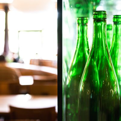 Grüne Glasflaschen in einer Vitrine. Unscharf im Hintergrund: Restaurantgäste sitzend am Tisch
