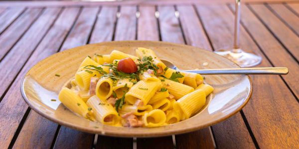 Rigatoni-Pasta mit Sahnesauce, Schinkenwürfeln, frischen Kräutern und einer Kirschtomate auf einem Holztisch.